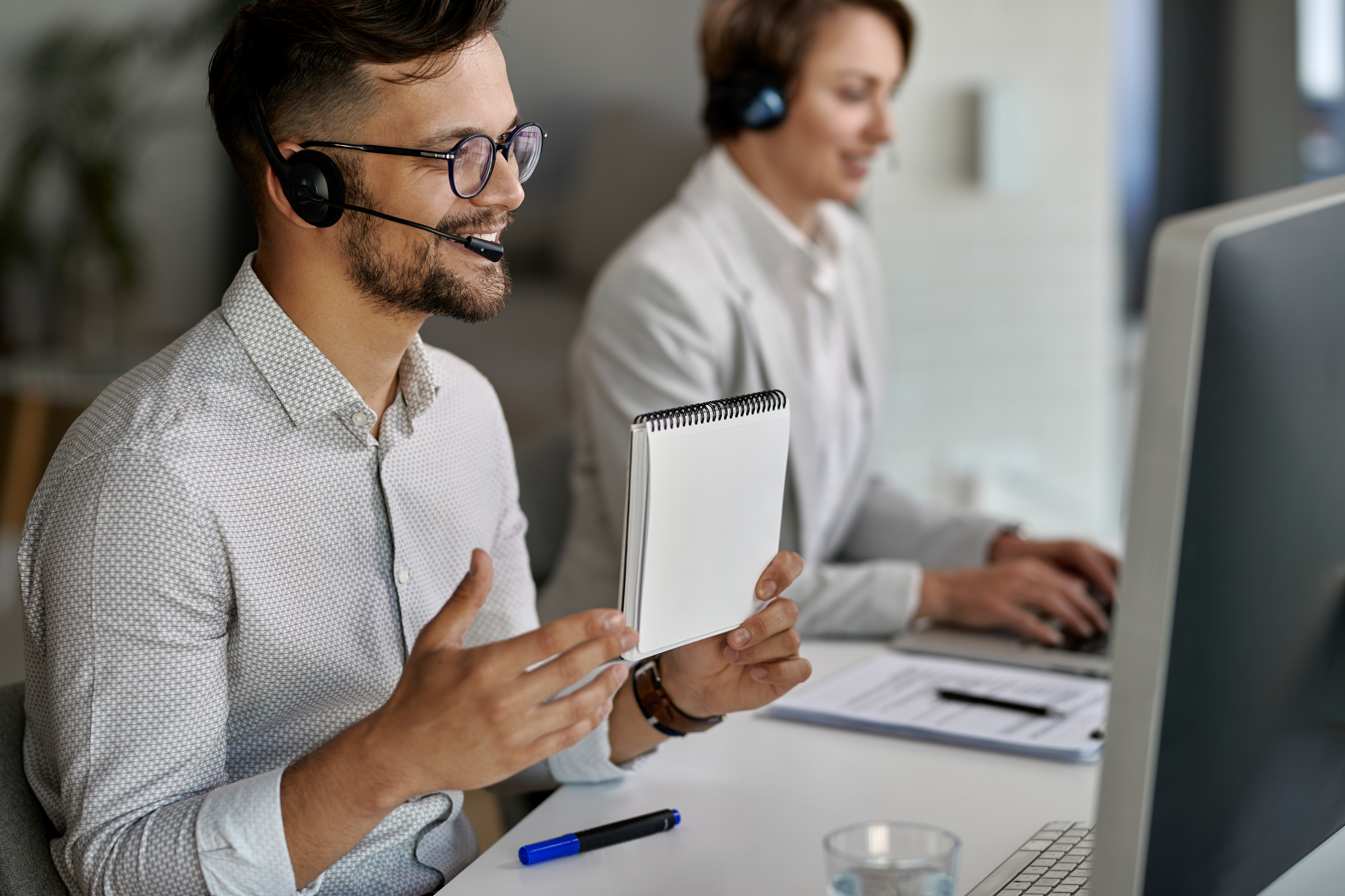 Customer Service Team at Work A male customer service representative wearing a headset and glasses, holding a notepad, while another representative works on a computer in the background.